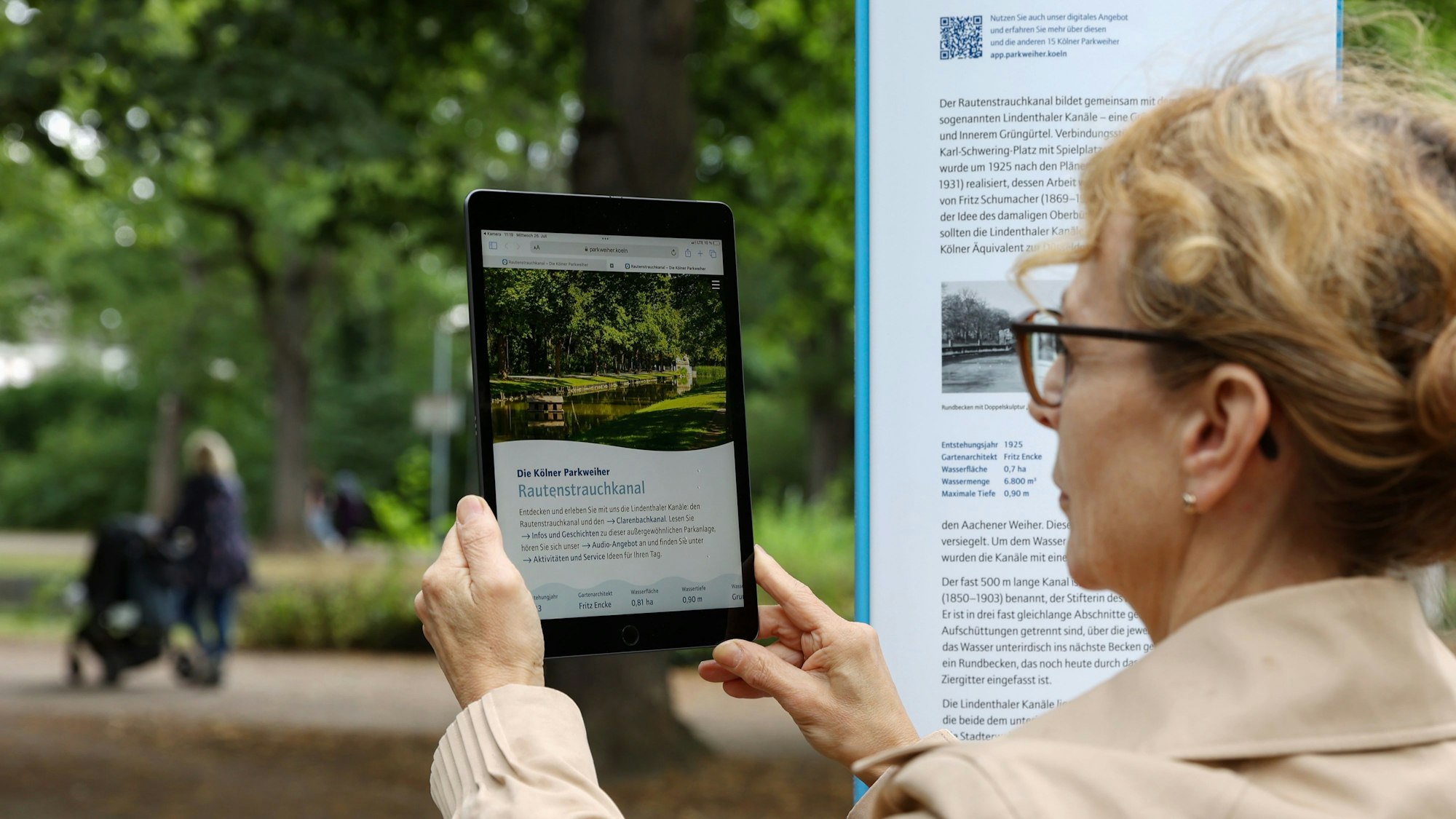 Eine Frau ruft Informationen über den Rautenstrauchkanal auf einem Tablet ab. Neben ihr steht eine neue Informationstafel.