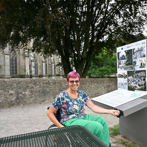Eine Frau sitzt im Rollstuhl und bedient lachend eine Kurbel, mit der eine Infostation am Altenberger Dom bedient wird.