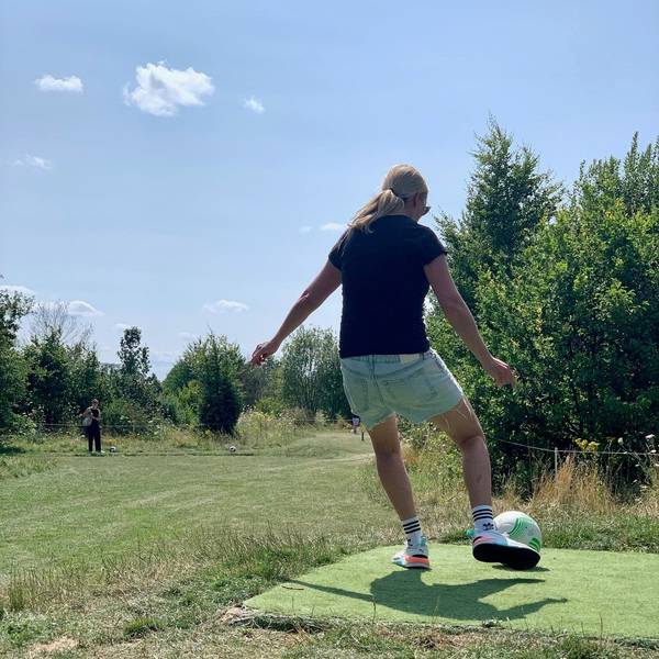 Eine Frau, die man von hinten sieht, schießt einen Fußball. Im Hintergrund sind Bäume und Wiese.