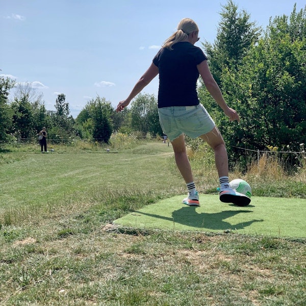 Eine Frau, die man von hinten sieht, schießt einen Fußball. Im Hintergrund sind Bäume und Wiese.