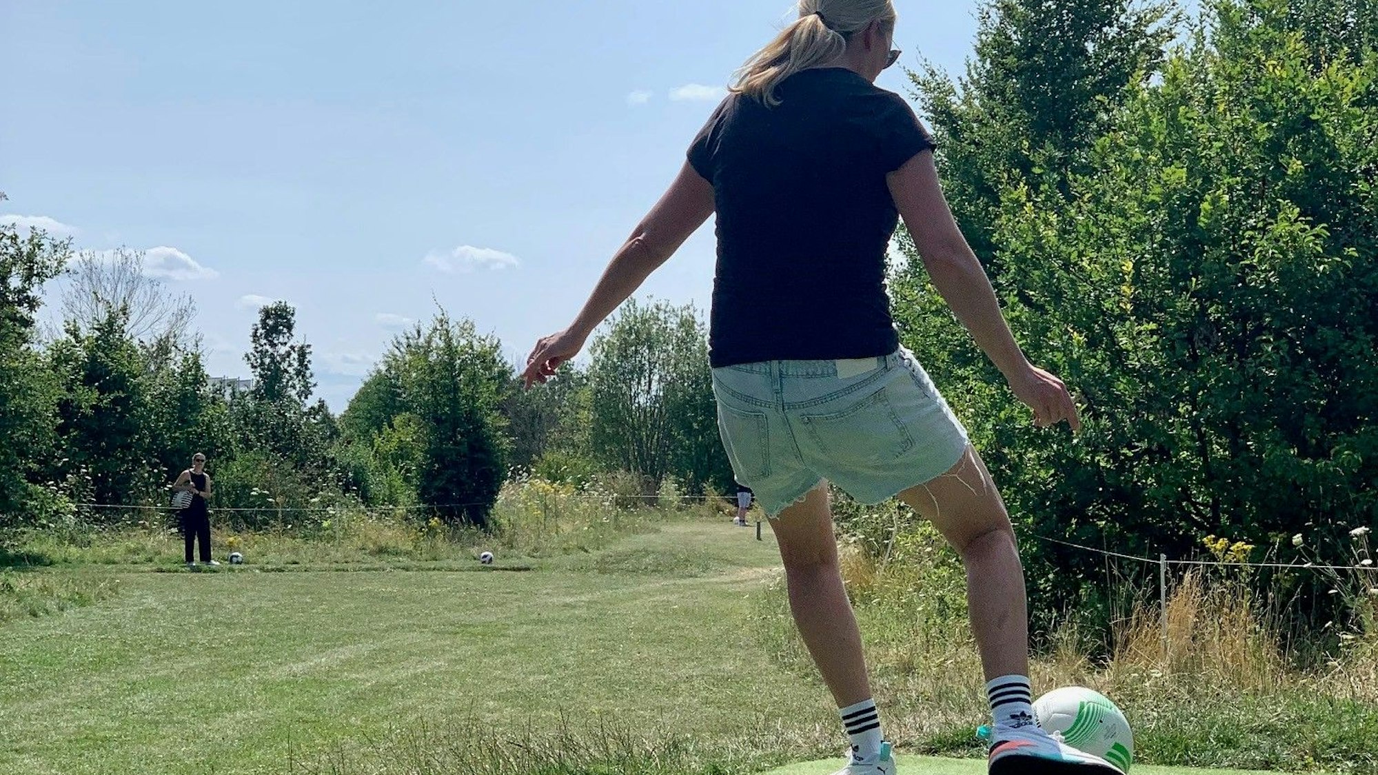 Eine Frau, die man von hinten sieht, schießt einen Fußball. Im Hintergrund sind Bäume und Wiese.
