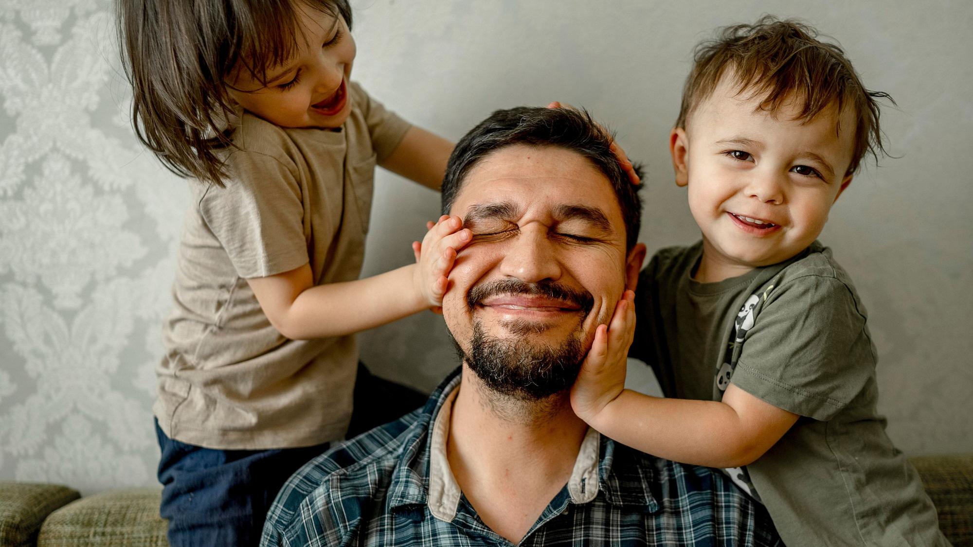 Ein Vater (mittig) sitzt mit seinen beiden Kindern (links und rechts) auf einem Sofa. Alle drei lächeln.
