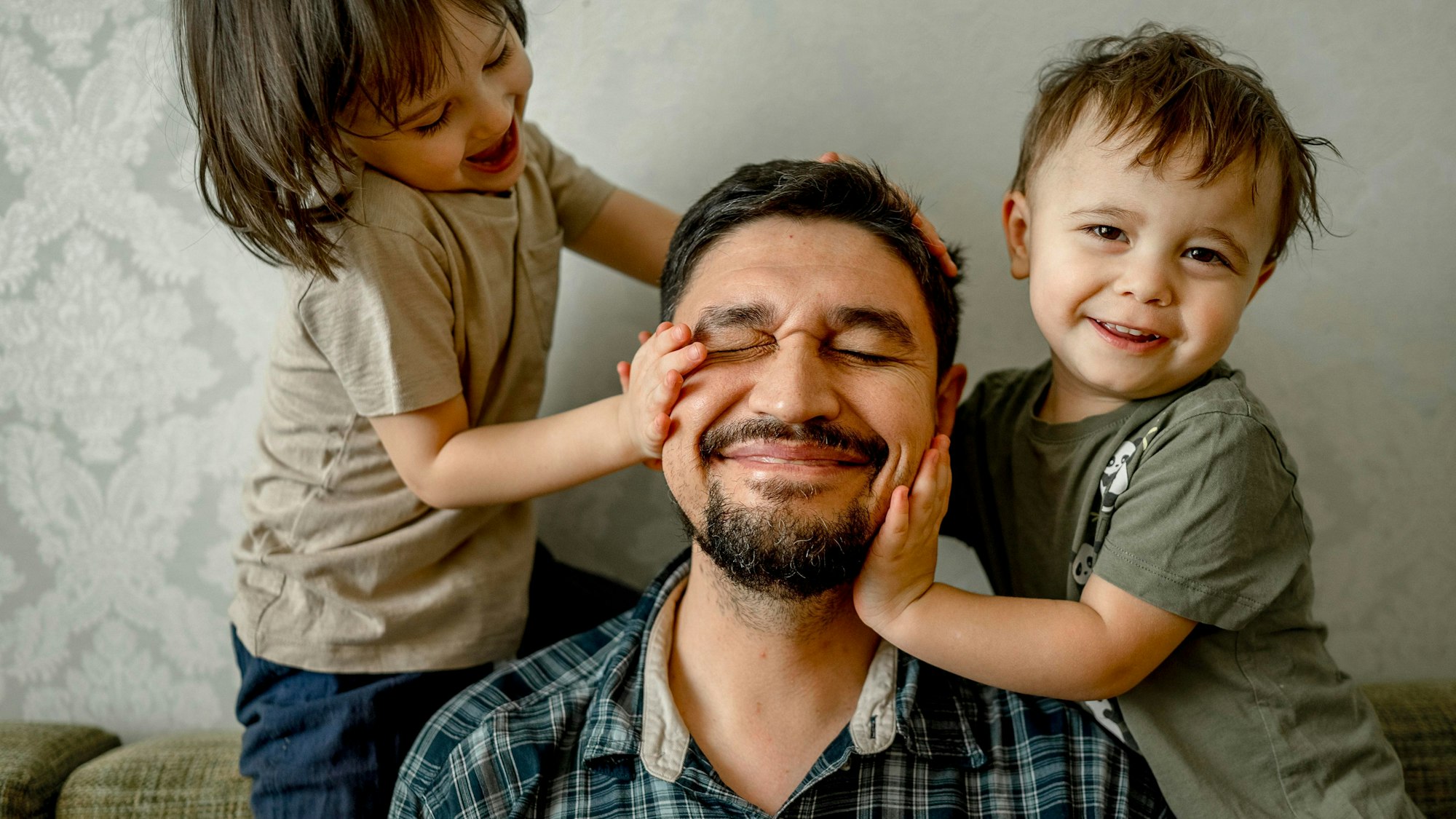 Ein Vater (mittig) sitzt mit seinen beiden Kindern (links und rechts) auf einem Sofa. Alle drei lächeln.