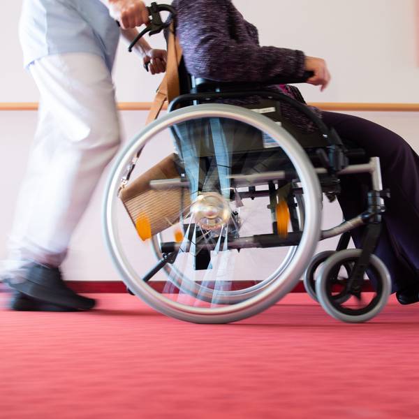 Ein Pfleger schiebt eine Bewohnerin eines Pflegeheims in einem Rollstuhl über den Flur.