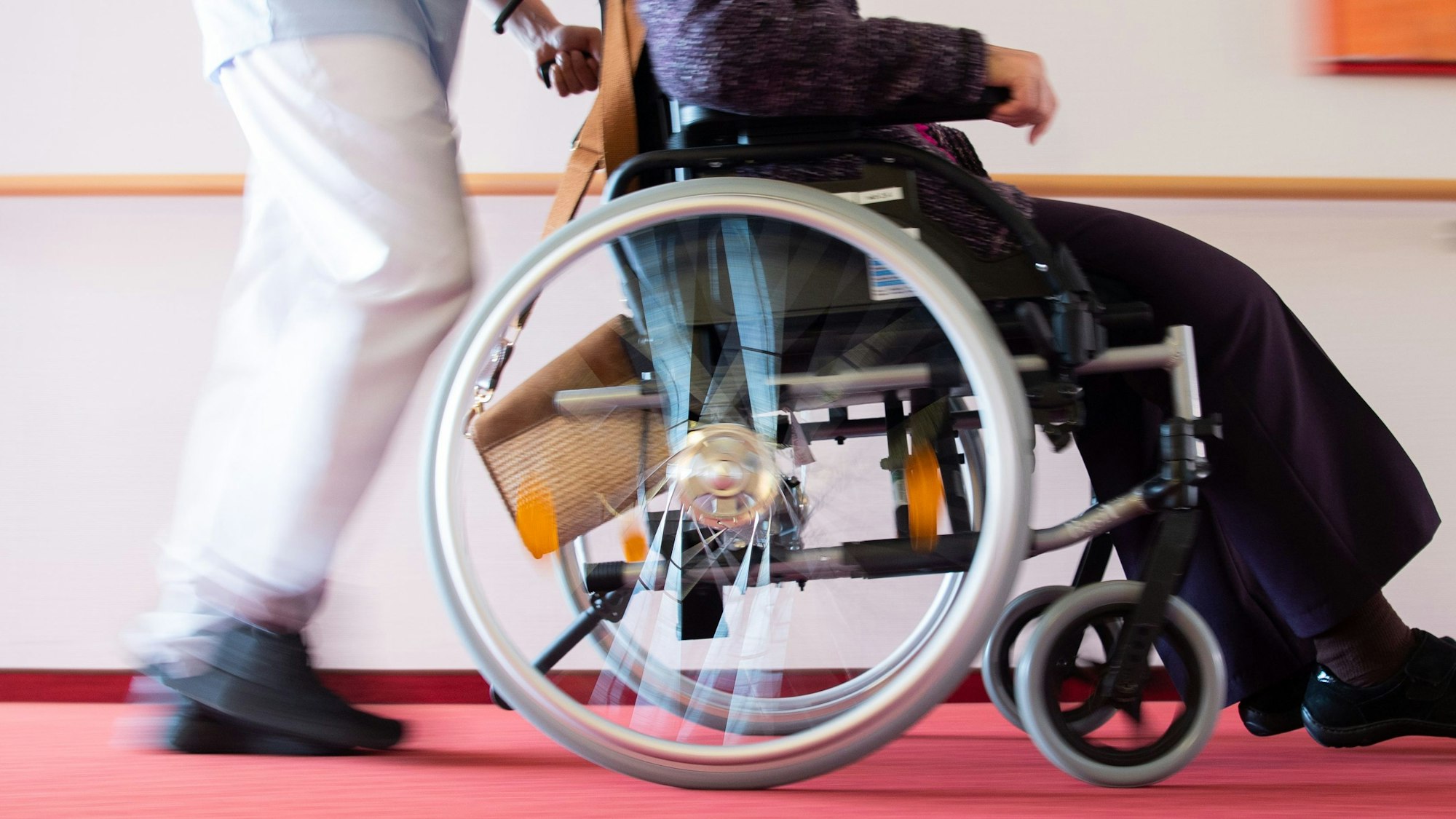 Ein Pfleger schiebt eine Bewohnerin eines Pflegeheims in einem Rollstuhl über den Flur.