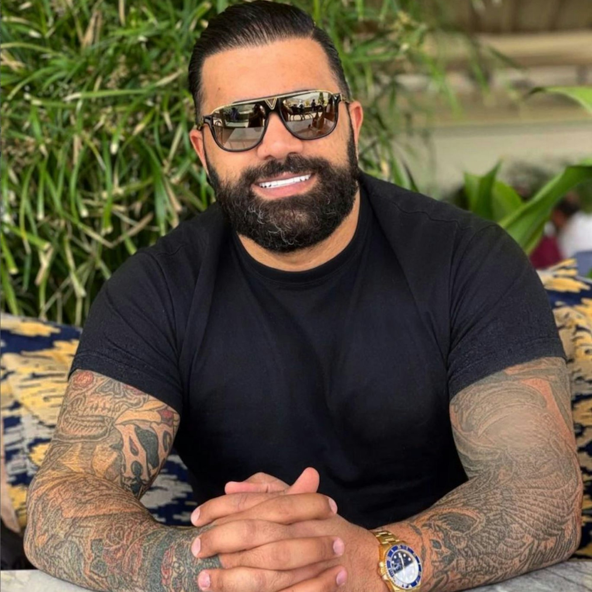 Ein Fahndungsfoto zeigt einen kräftigen Mann mit schwarzem Bart und Sonnenbrille. Er trägt ein schwarzes T-Shirt, eine goldene Uhr und hat tätowierte Arme.