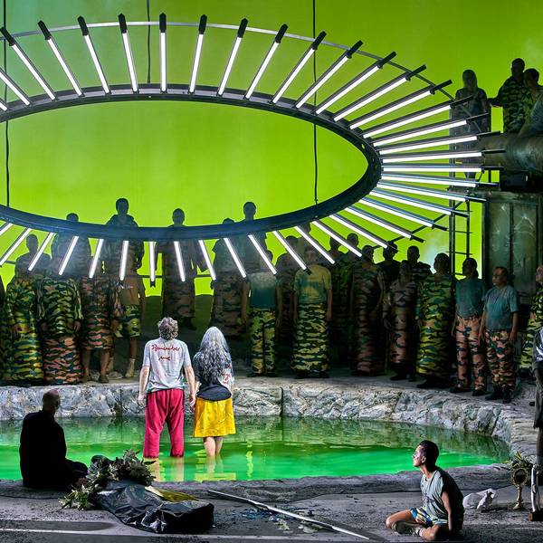 Auf der Bühne stehen Parsifal und Kundry in einer Art Teich, über ihnen ein großer Kreis mit LED-Lampen. Um sie herum stehen zahlreiche Menschen.