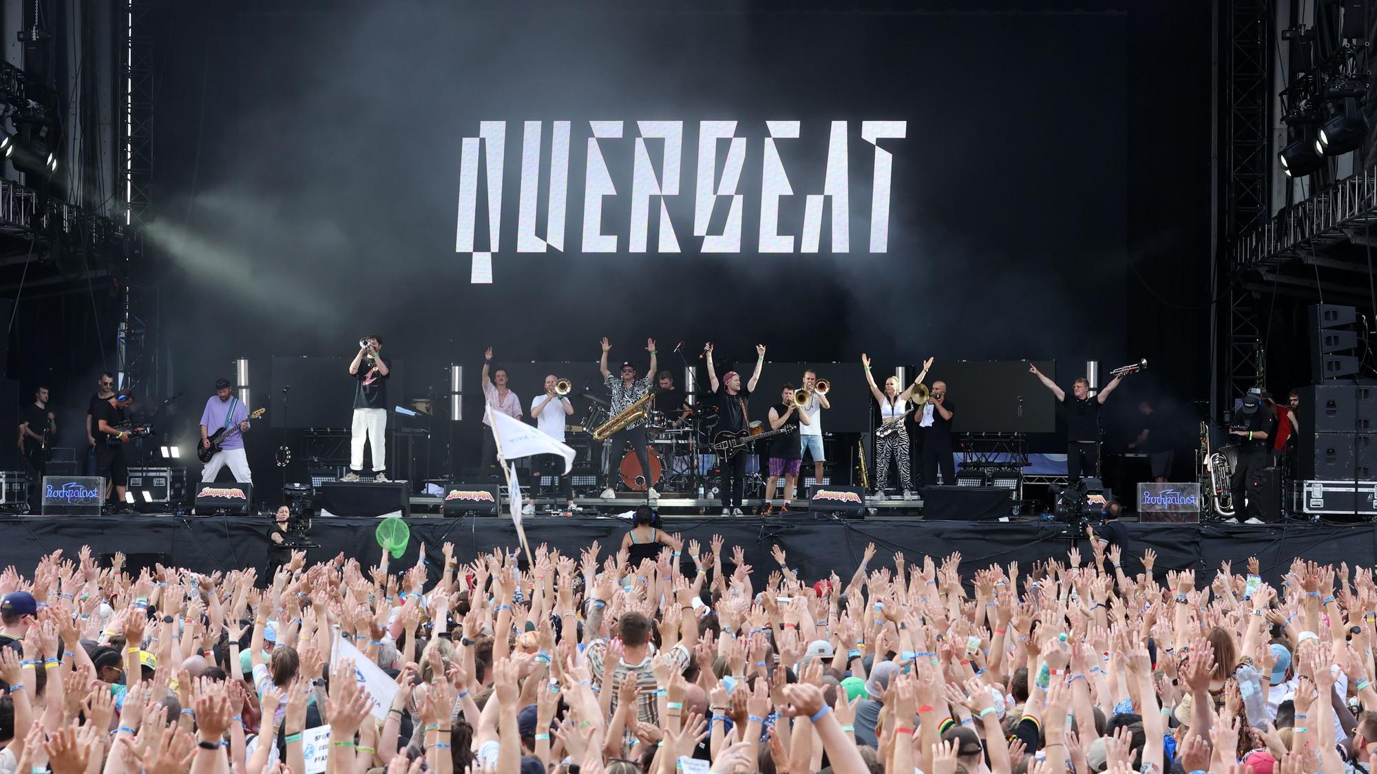 Hunderte Menschen stehen vor einer Bühne, mit den Händen in der Höhe. Auf der Bühne stehen mehrere Personen. Dort ist „Querbeat “zu lesen.