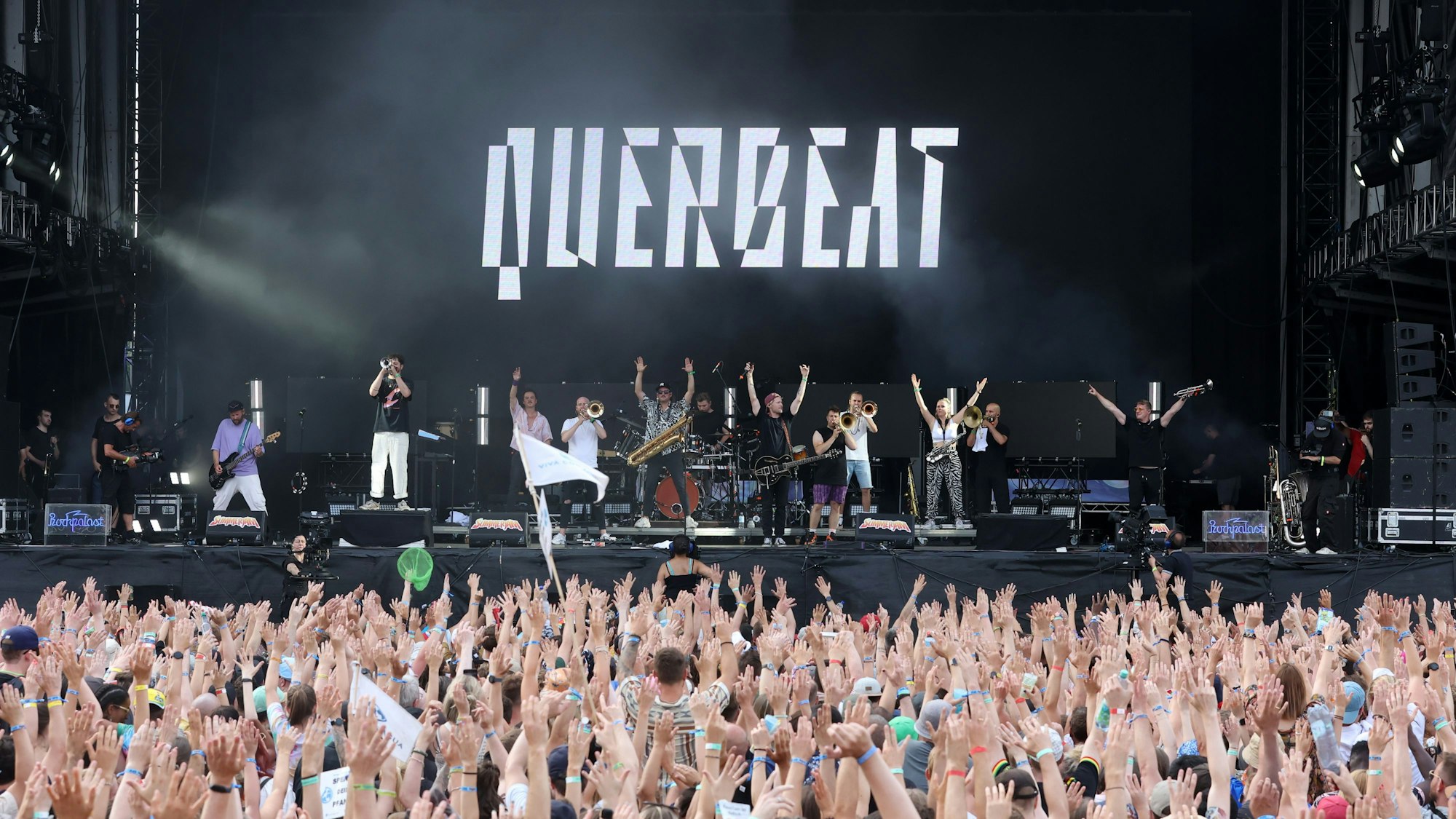 Jubelnde Menge vor einer Bühne, auf der eine Band steht. Im Hintergrund der Schrifzug „Querbeat“ zu sehen