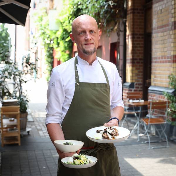 Mann in Kochkleidung steht vor einem Restaurant und hält drei Teller in der Hand
