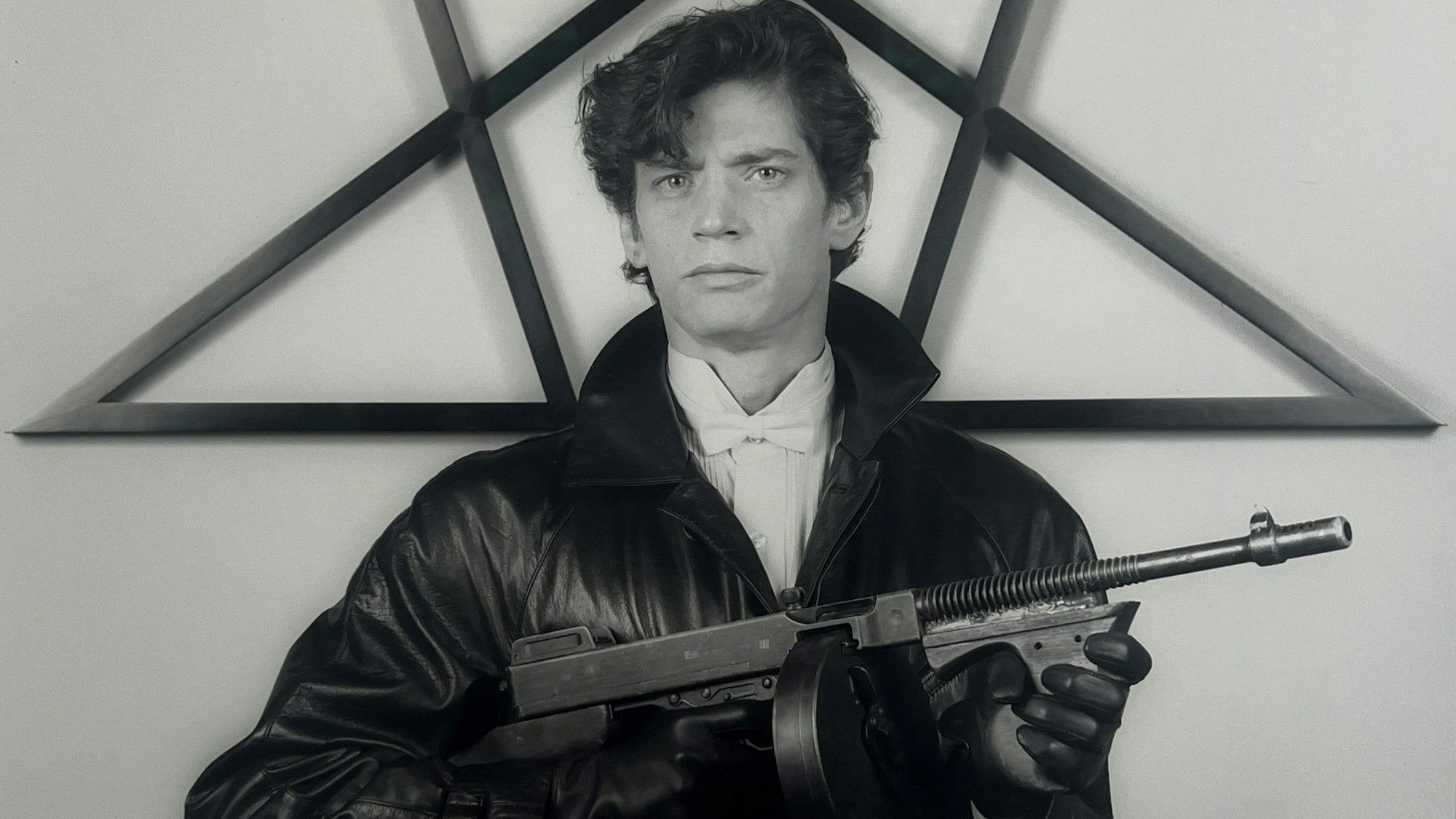 Der Fotograf Robert Mapplethorpe posiert mit Maschinenpistole vor schwarzem Stern. Er trägt eine schwarze Lederjacke.