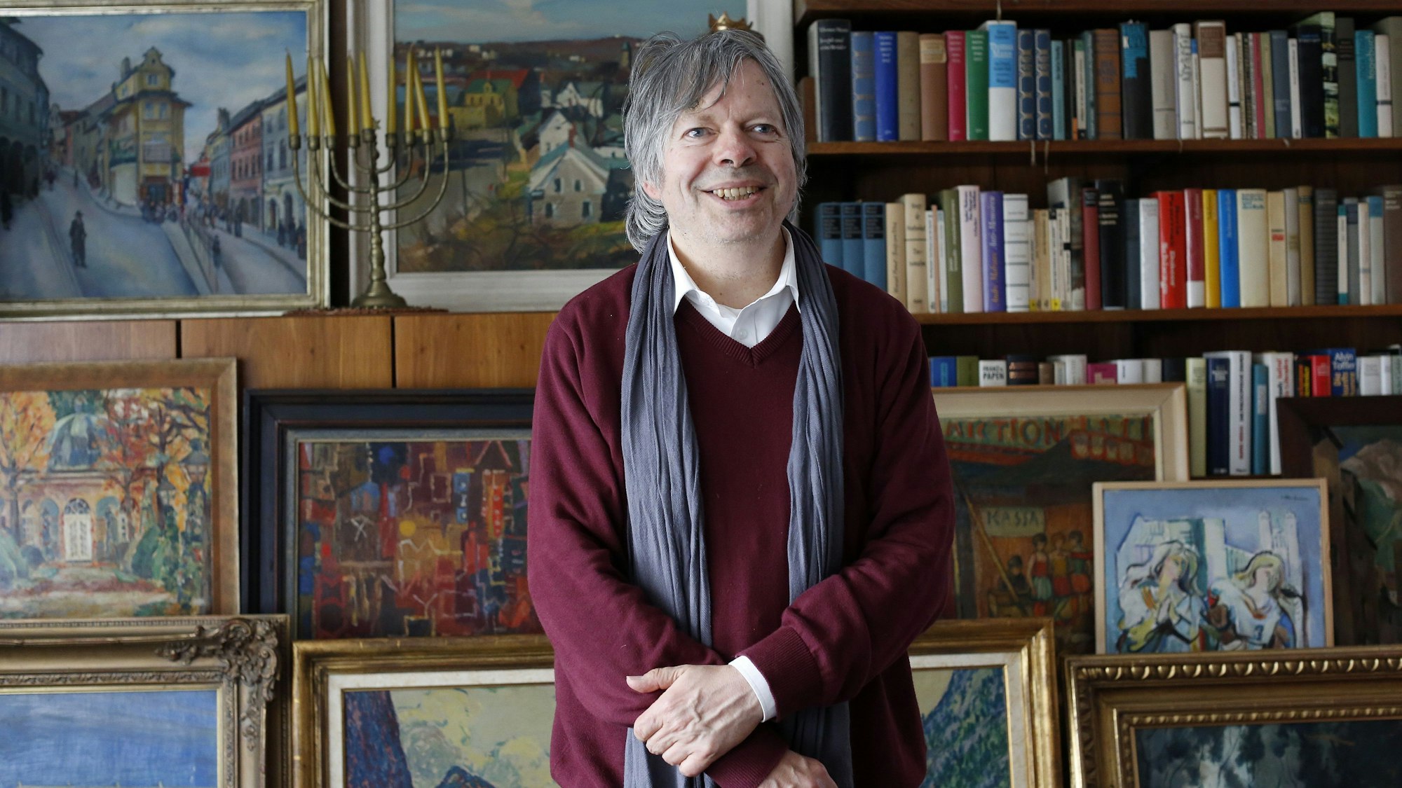 Das Foto zeigt den Verleger in seinem Arbeitszimmer, hinter ihm hängen Bilder an der Wand, vor einem Bücherregal stehen weitere Motive.