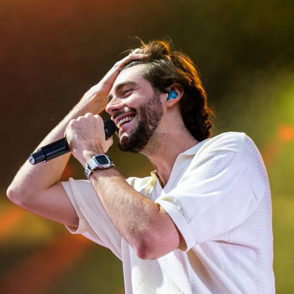 Alvaro Soler hat braune Haare und einen Bart. Er trägt ein weißes Hemd und hat am Handgelenk.eine graue Uhr. Er hält ein Mikrofon und kämmt sich mit der Hand die Haare nach hinten.
