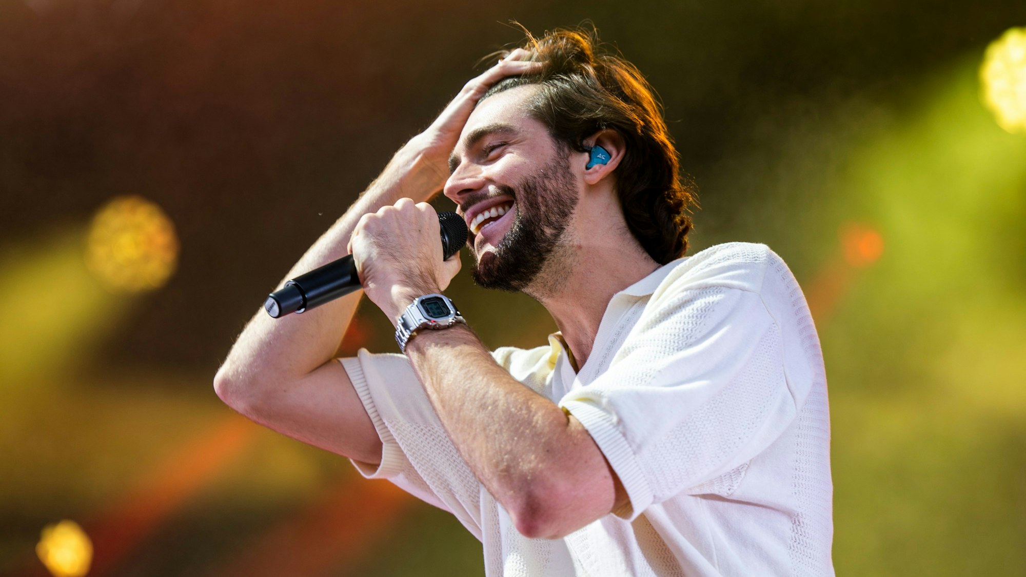 Alvaro Soler hat braune Haare und einen Bart. Er trägt ein weißes Hemd und hat am Handgelenk.eine graue Uhr. Er hält ein Mikrofon und kämmt sich mit der Hand die Haare nach hinten.