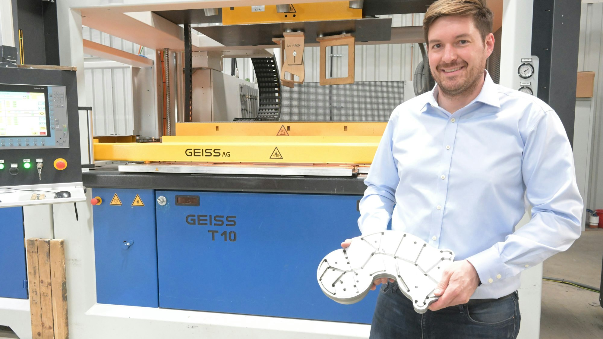 Constantin Fischer steht vor einer Thermoform-Maschine und hält in der Hand ein Werkzeug zur Weiterverarbeitung des gerade geformten Plastikproduktes.