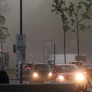 Mehrere Autos fahren mit Scheinwerferlicht durch den Regen in der Kölner Südstadt. Im Hintergrund reißen Sturmböen an den Bäumen. (Symbolbild)