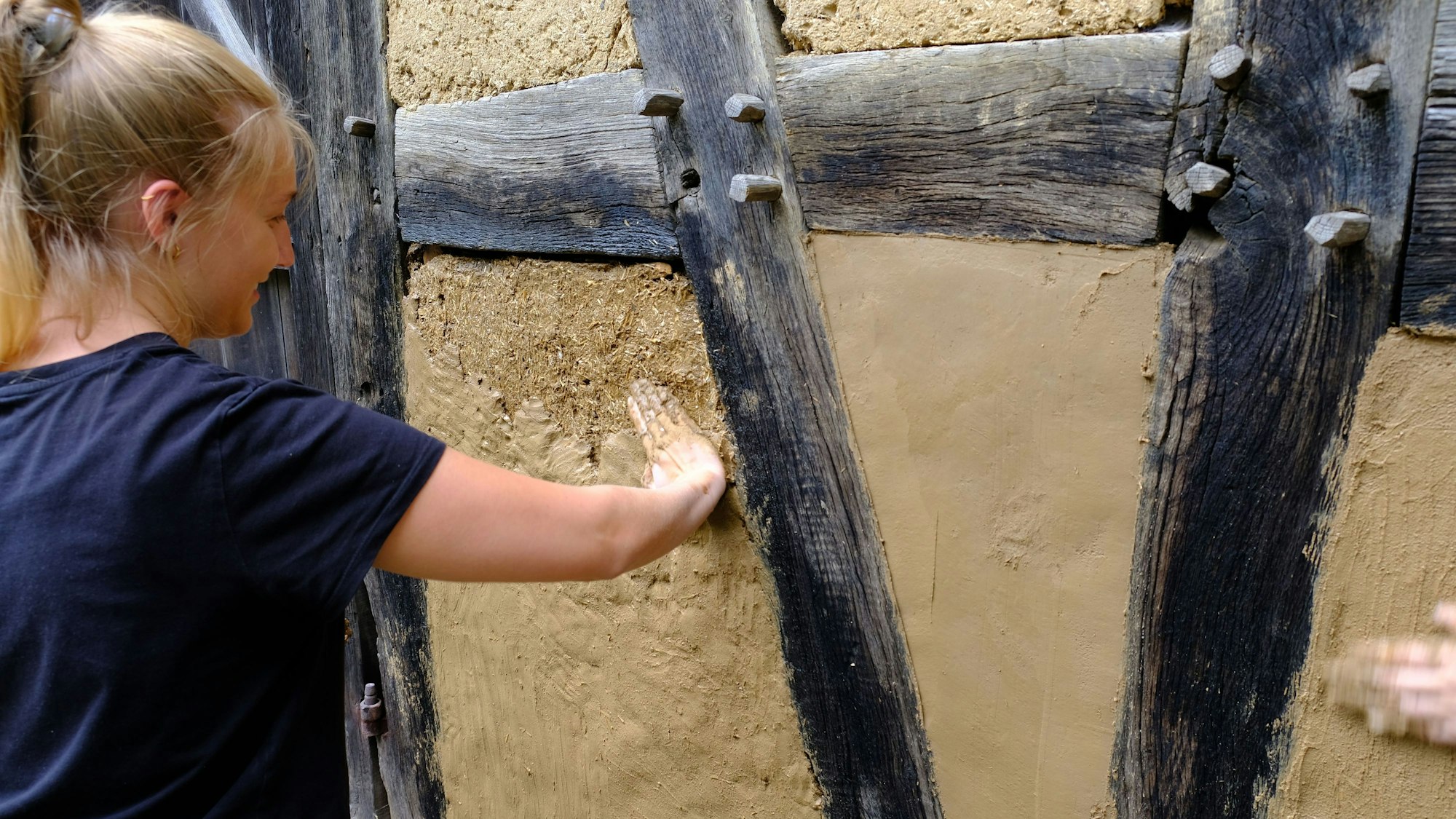 Eine junge Frau glättet den aufgetragenen Lehm auf einer Fachwerkwand mit der Hand.