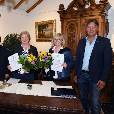 Mehrere Personen stehen in einem Trauzimmer. Zwei Frauen halten Zertifikate in den Händen.