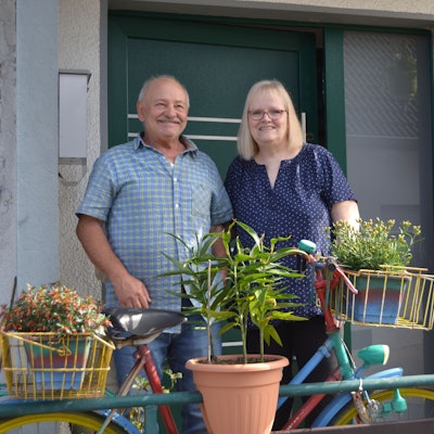 Ein Ehepaar steht vor einer Haustüre. Vor ihnen steht ein bepflanztes Fahrrad.
