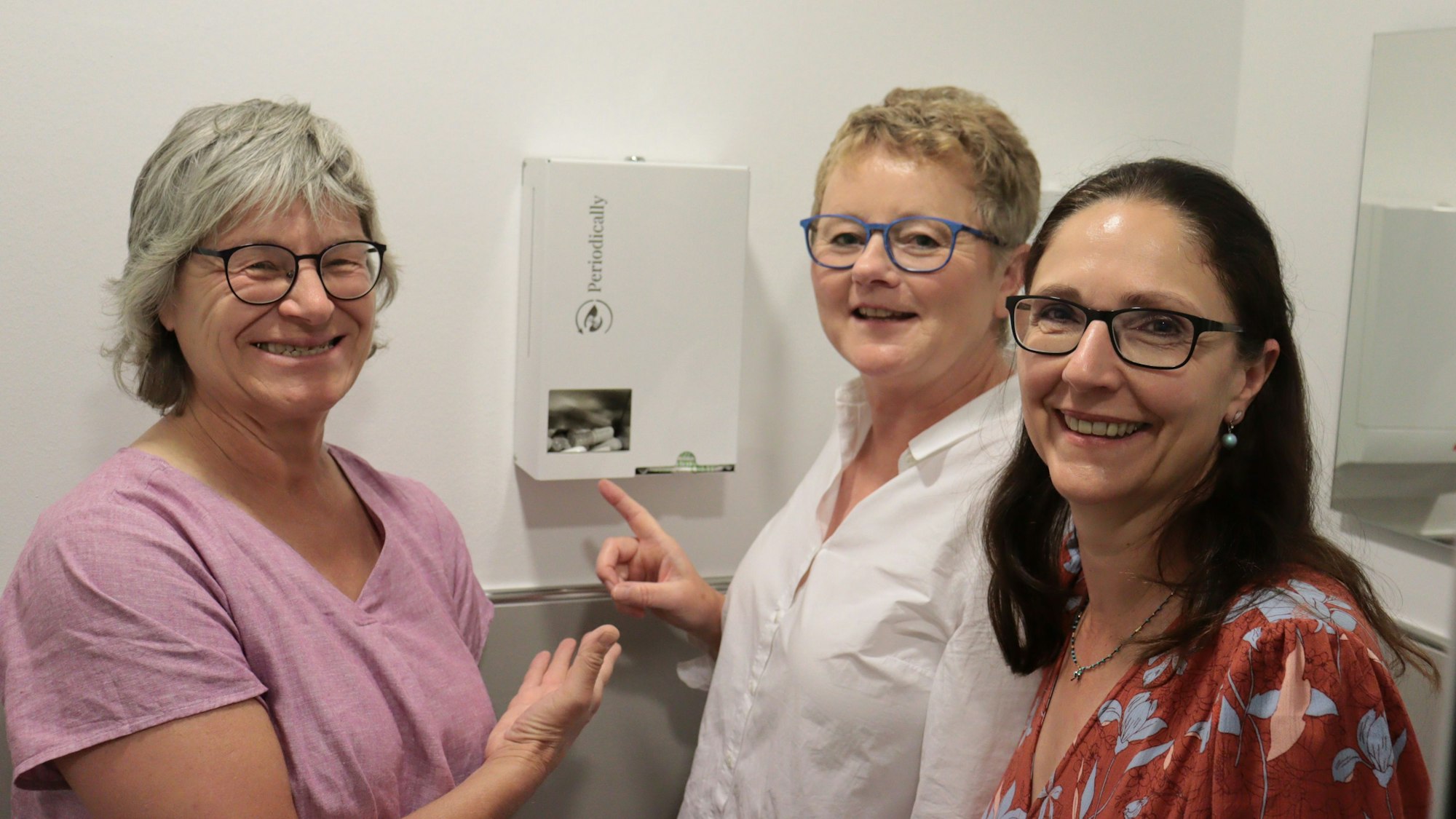 Drei Frauen präsentieren einen Spender für kostenlose Menstruationsartikel.