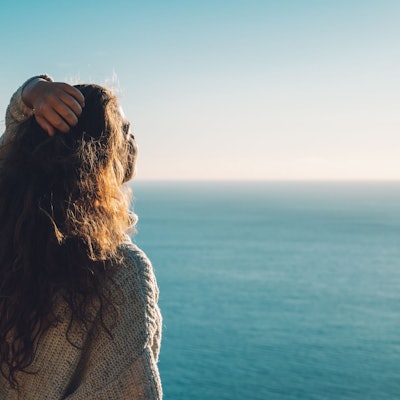 Eine Frau steht am Meer und streckt den Kopf Richtung Sonne.