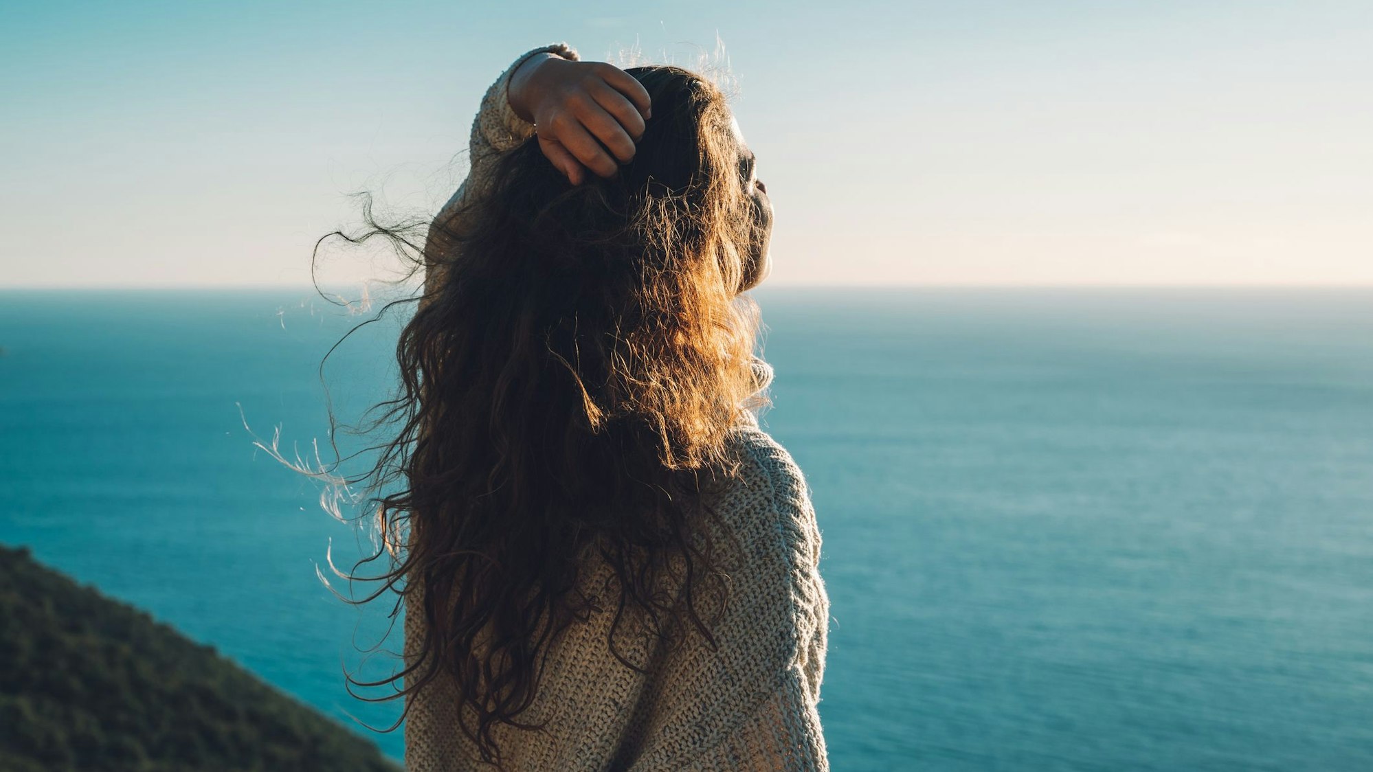 Eine Frau steht am Meer und streckt den Kopf Richtung Sonne.
