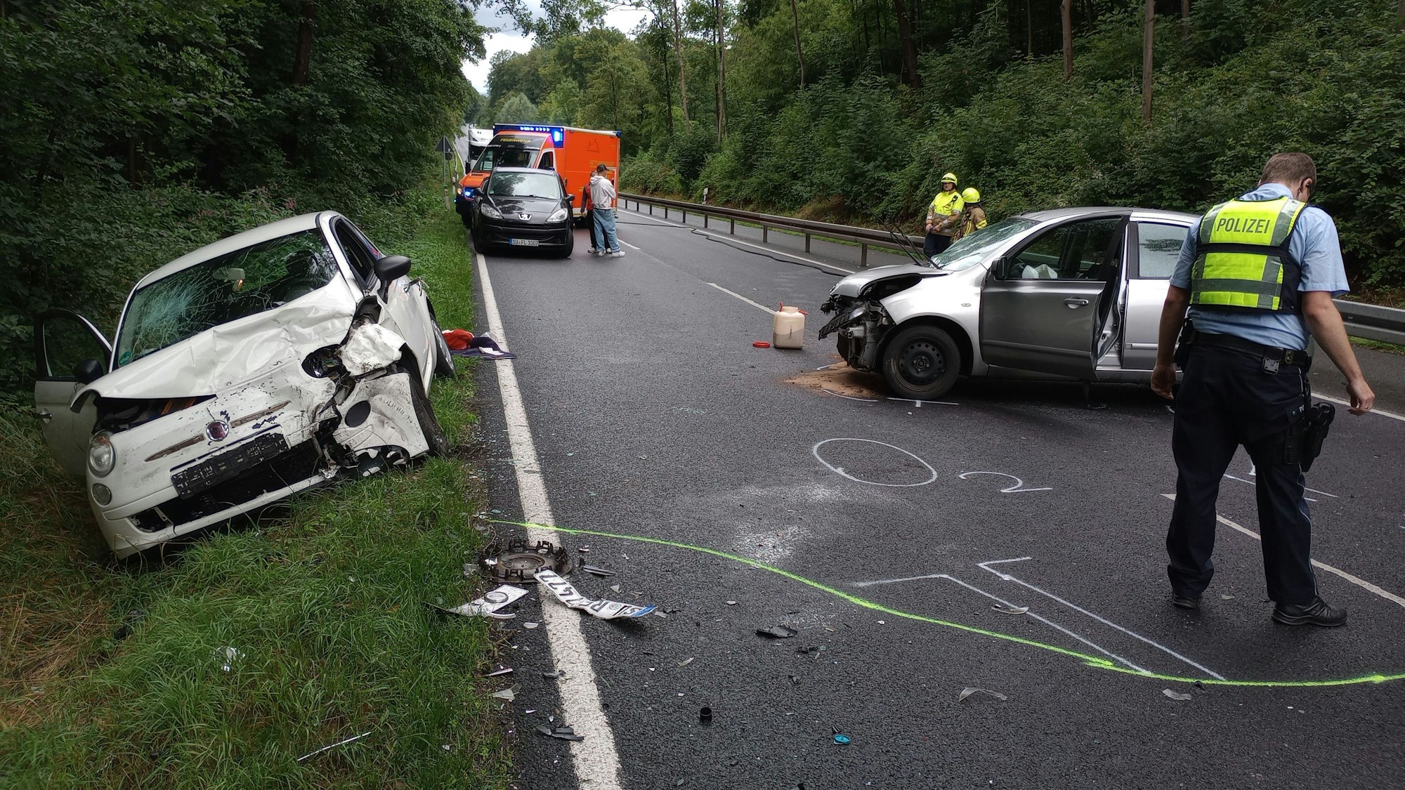Zwei stark beschädigte Autos stehen auf der B56 bei Siegburg auf der Straße. Polizisten stehen um die Unfallstelle herum. Im Hintergrund steht ein Krankenwagen mit Blaulicht.