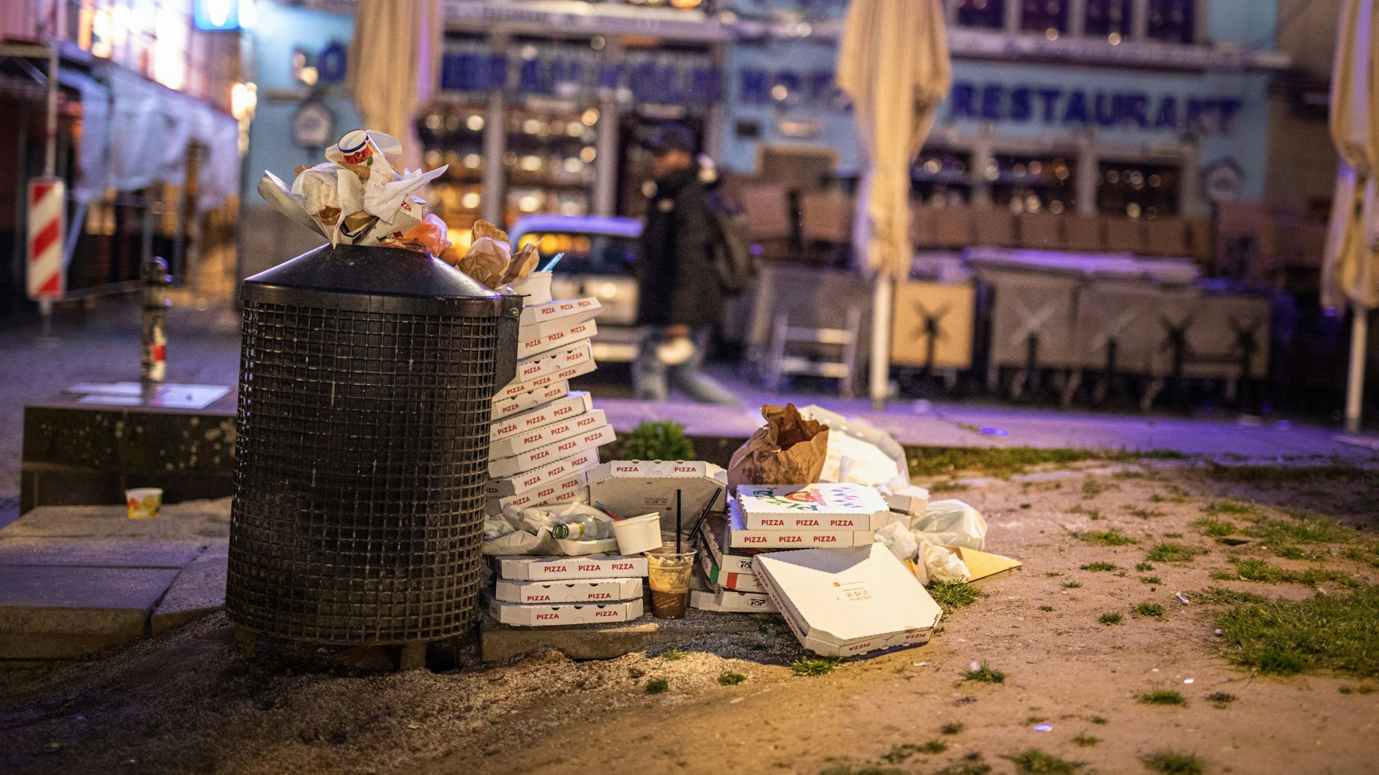 Müll stapelt sich neben einem Mülleimer. Zahlreiche Pizzakartons liegen weiträumig um den Müllbehälter verstreut. Im Bildhintergrund ist die Fassade eines Restaurants in der Kölner Altstadt zu sehen.