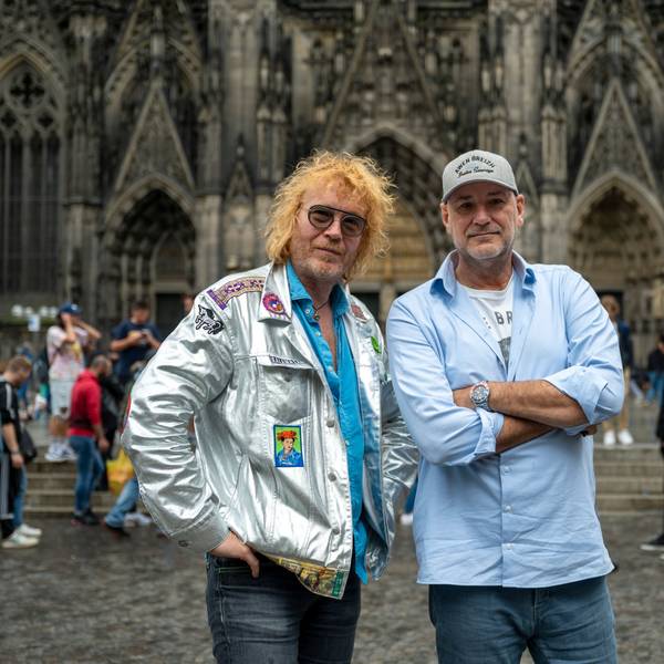 Christof Stein-Schneider in silbener Jacke und Kai Wingenfelder im blauen Hemd und Kappe, posieren vor dem Kölner Dom.