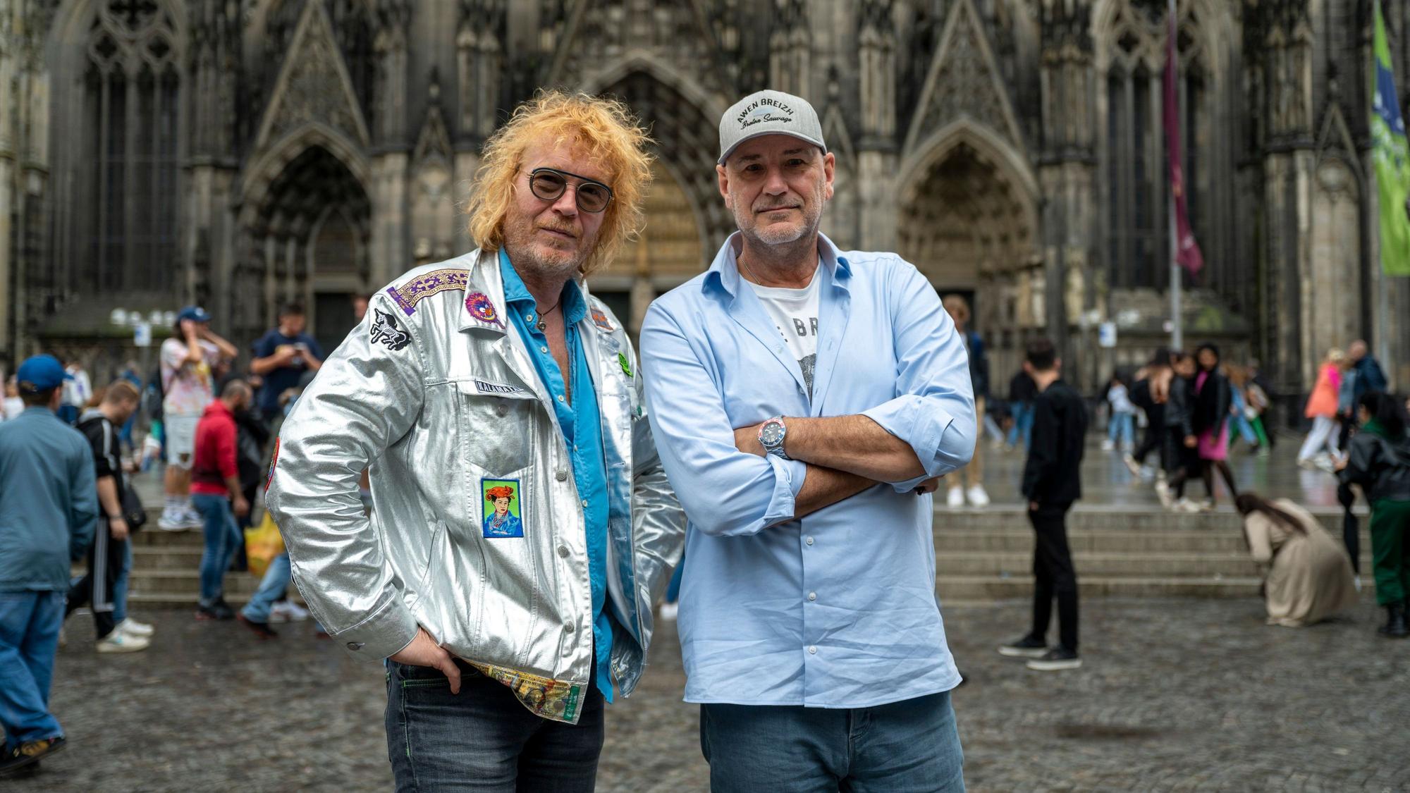 Christof Stein-Schneider in silbener Jacke und Kai Wingenfelder im blauen Hemd und Kappe, posieren vor dem Kölner Dom.