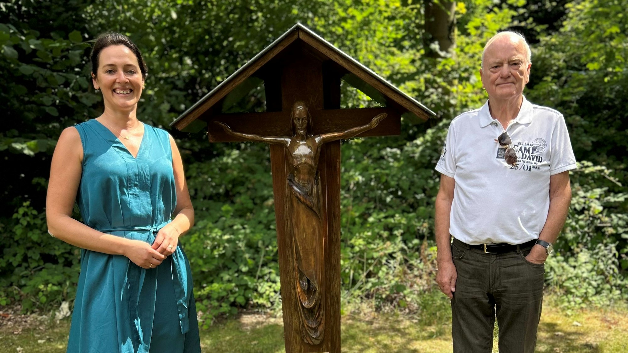 Eine Frau und ein Mann stehen auf einem Friedhof neben einem Holzkreuz.