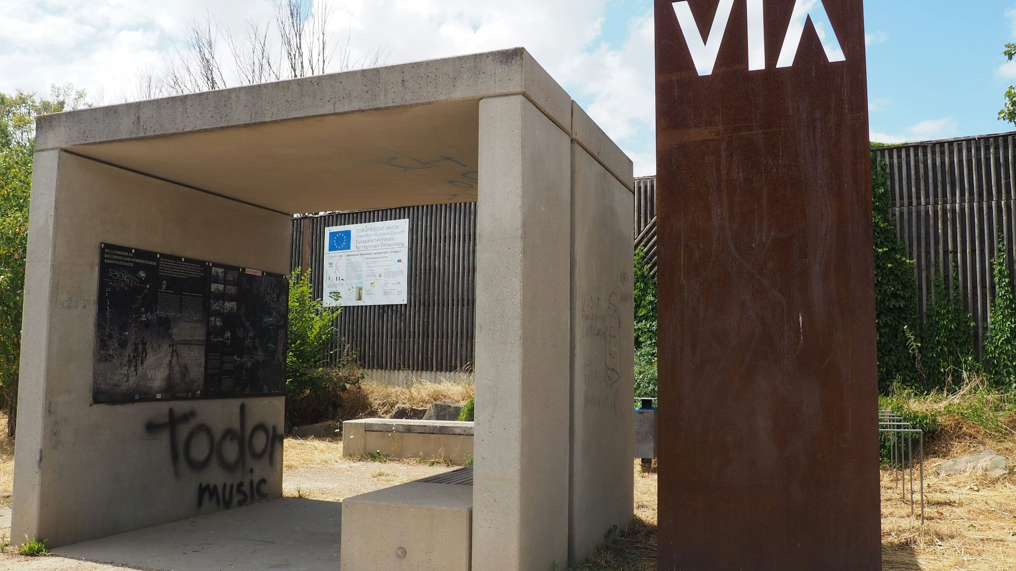 Ein offenes Bauwerk aus Beton spendet Schatten, daneben steht eine Stele aus verrostetem Metall.