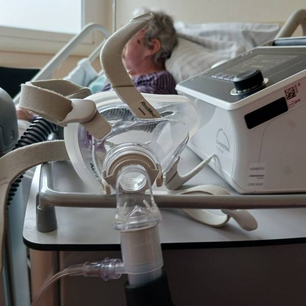 Eine Seniorin liegt in einem Bett im Krankenhaus. Vor ihr steht ein Atemgerät.