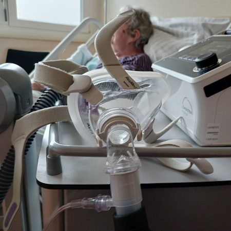 Eine Seniorin liegt in einem Bett im Krankenhaus. Vor ihr steht ein Atemgerät.