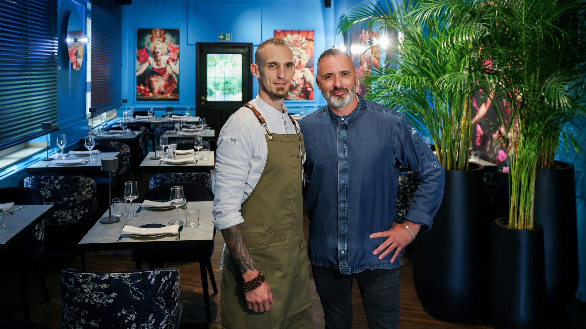 Dias und Becker stehen in der Weinbar, die Wände sind blau.