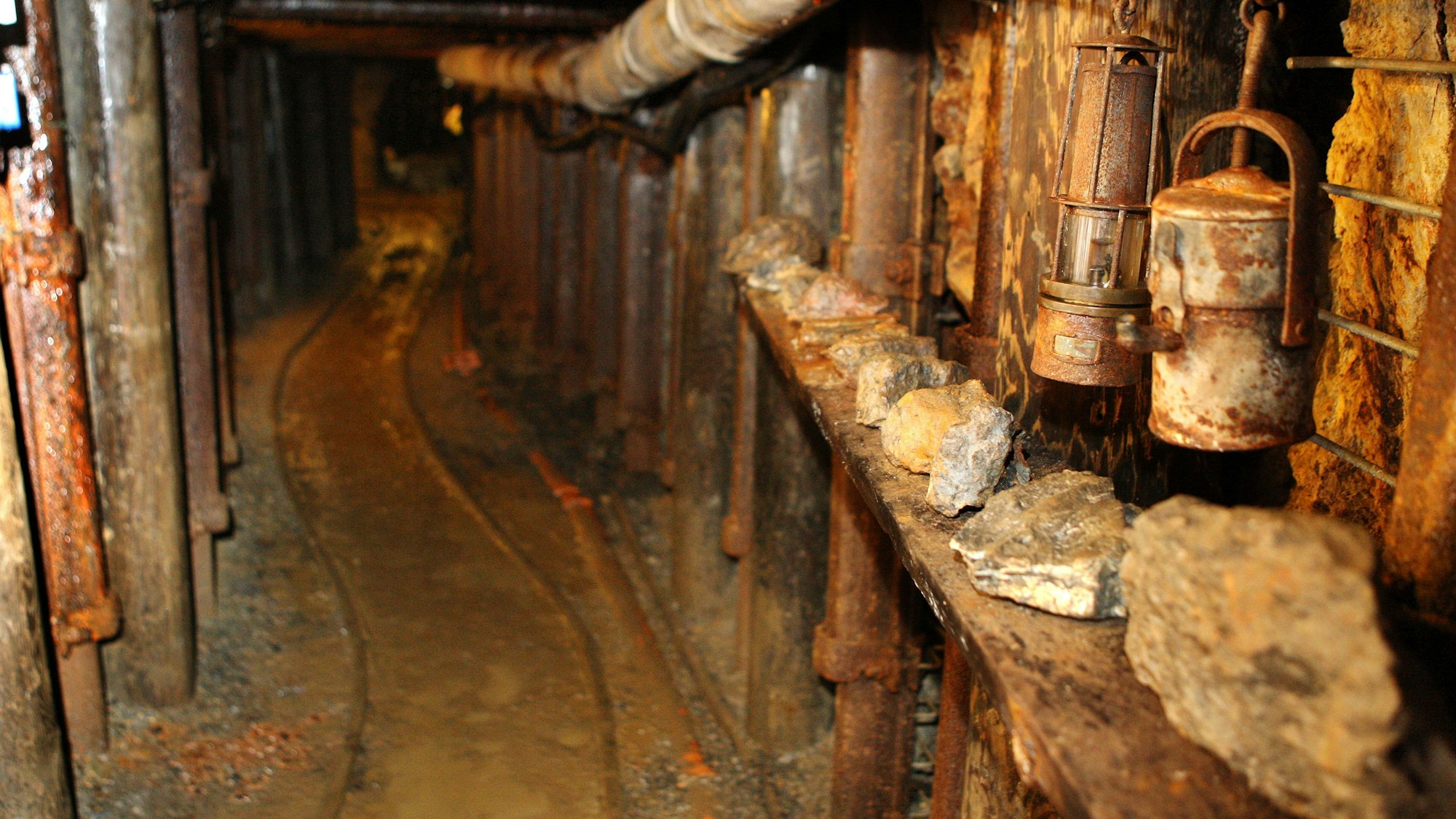 Ein Stollen mit auf dem Boden verlegten Schienen führt in einen Berg, an der Wand hängen Bergwerkslampen.