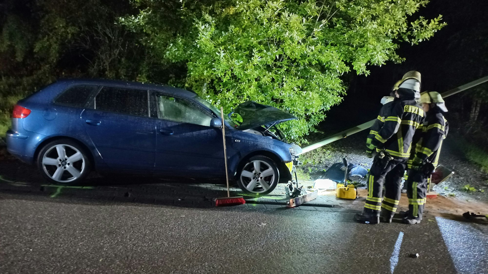 Ein Auto steht vor einer umgeknickten Straßenlampe, daneben drei Feuerwehrleute.