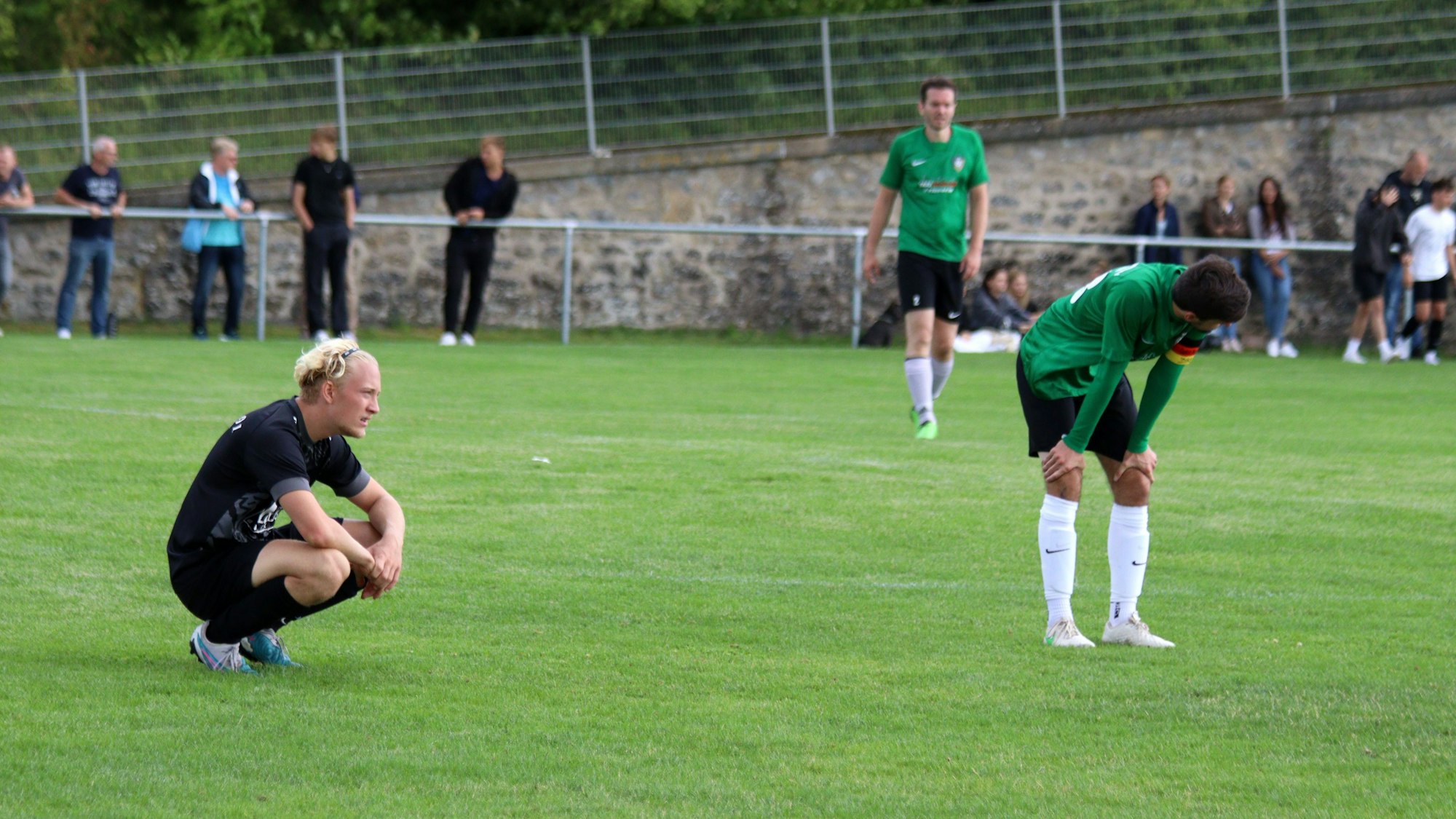 Erft-Spieler Luc-Seal Roggendorf hockt erschöpft auf dem Rasen, daneben steht FlaKis Tom Neukirch, die Hände auf die Knie abgestützt.