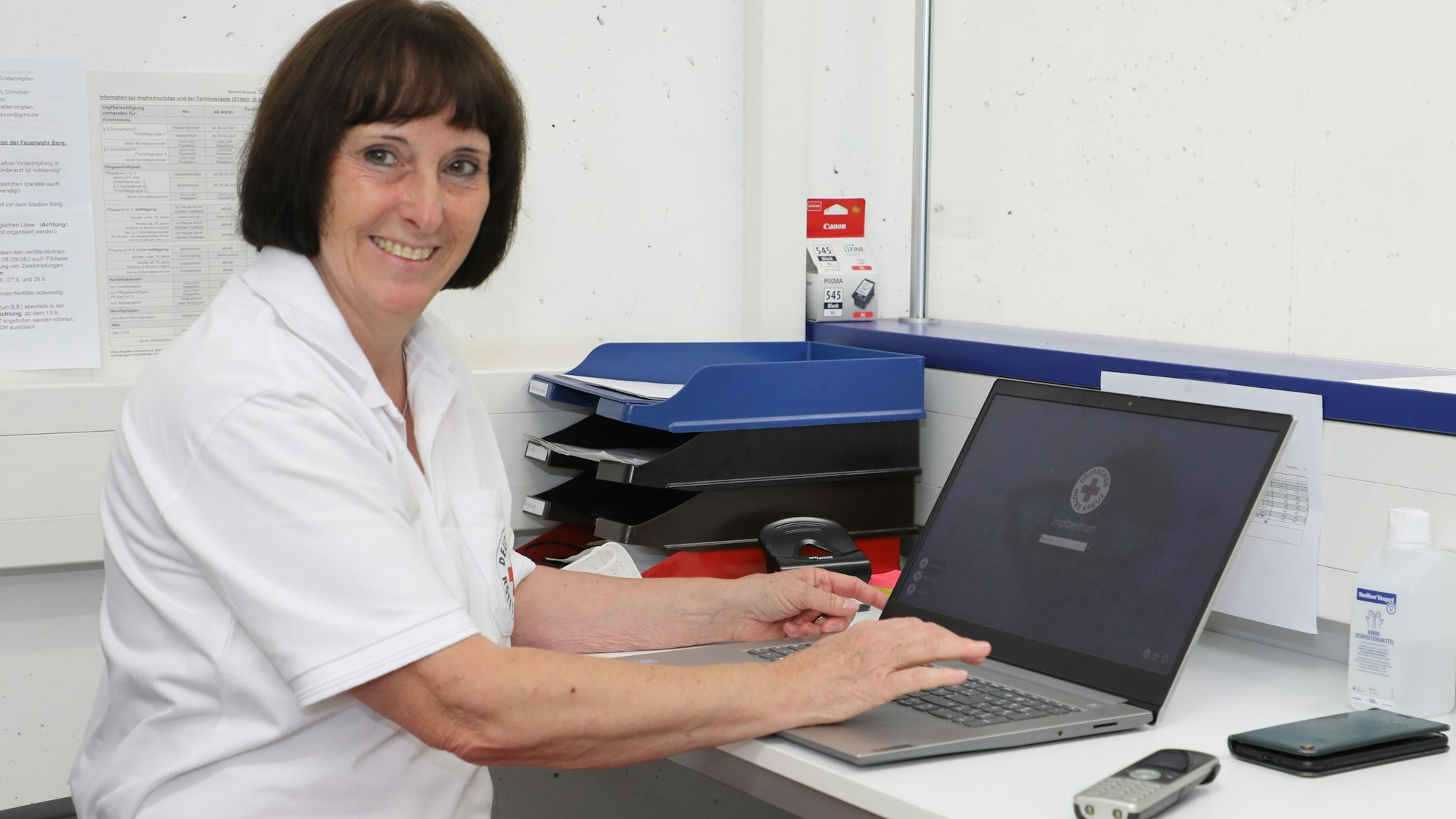 DRK-Kreis- und -Ortsvorsitzende Ingeborg Schmidt sitzt vor einem Laptop an einem Schreibtisch, auf dem auch ein Handy und ein Telefon liegen.