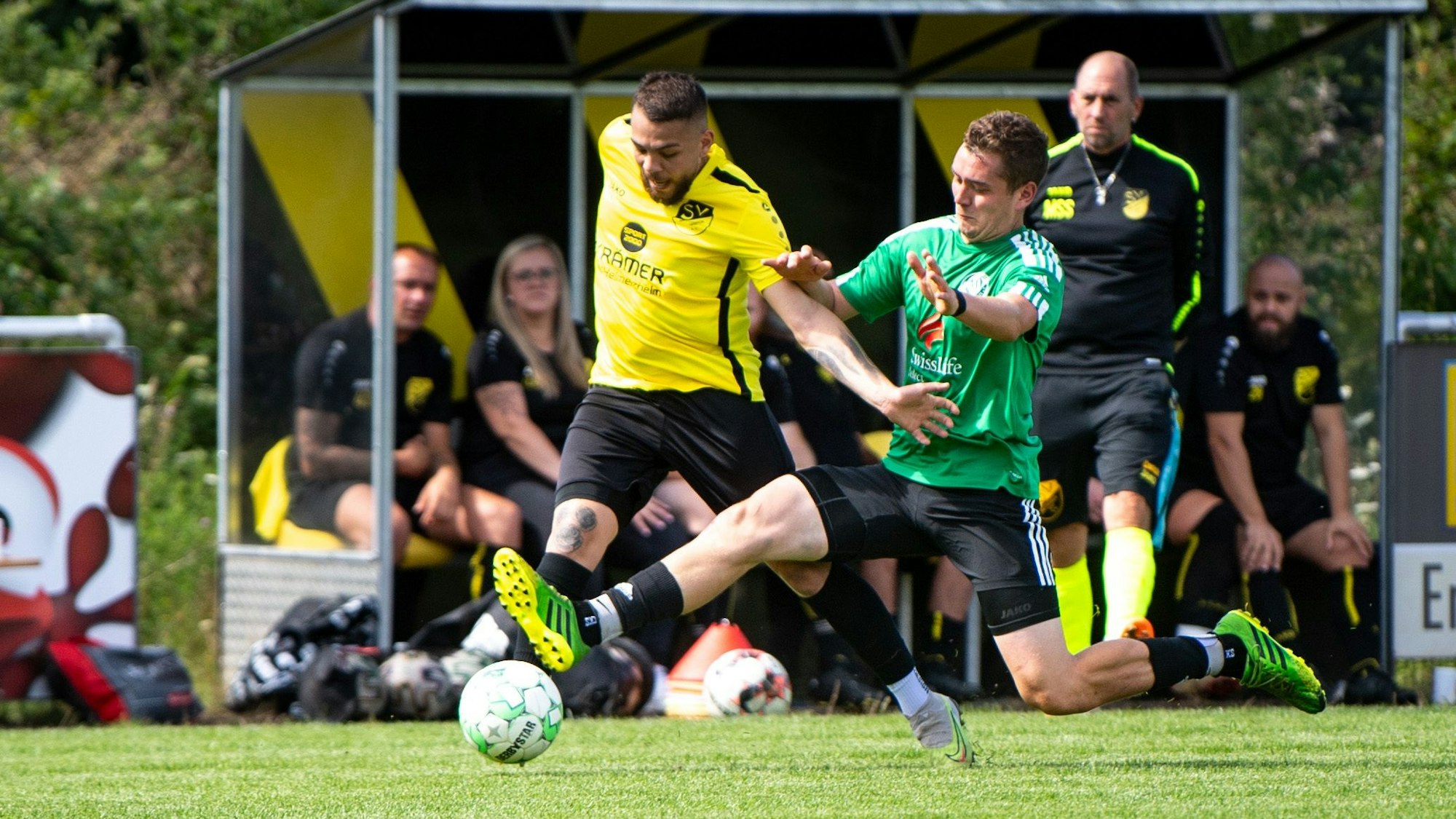 Zweikampf zwischen zwei Spielern von Metternich und Schöneseiffen, im Hintergrund die Metternicher Trainerbank.