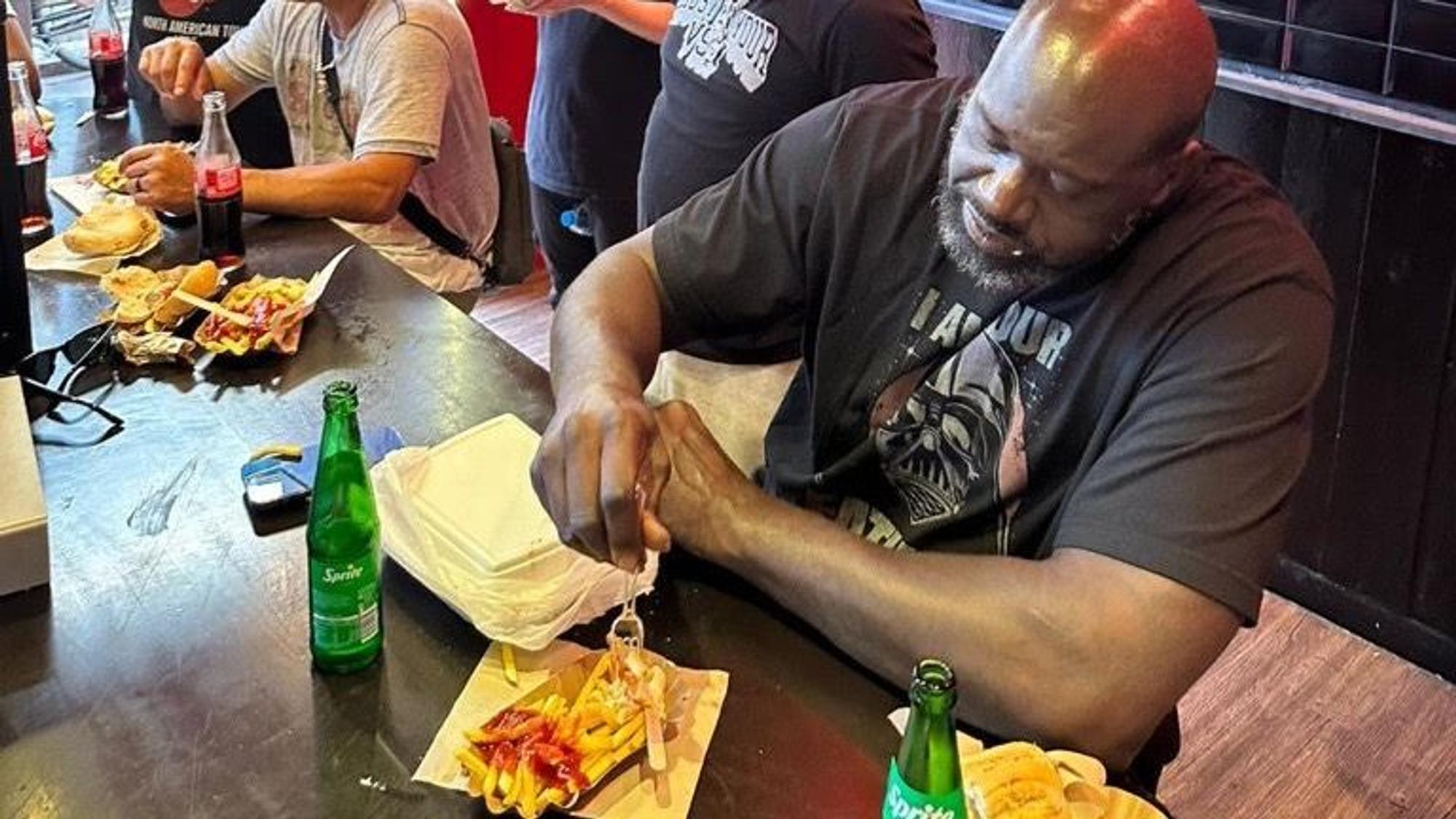 Shaquille O'Neal isst am 28. Juli 2023 im Weltmeister am Kölner Ebertplatz eine Pommes mit Wurst.