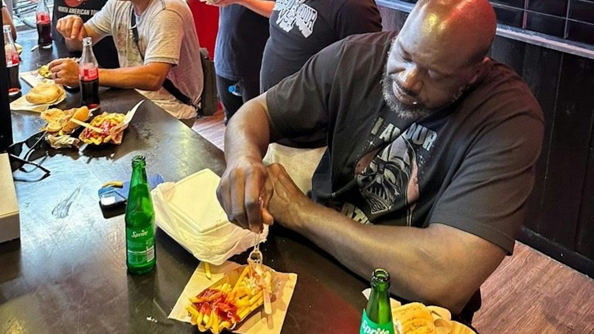 Shaquille O'Neal isst am 28. Juli 2023 im Weltmeister am Kölner Ebertplatz eine Pommes mit Wurst.