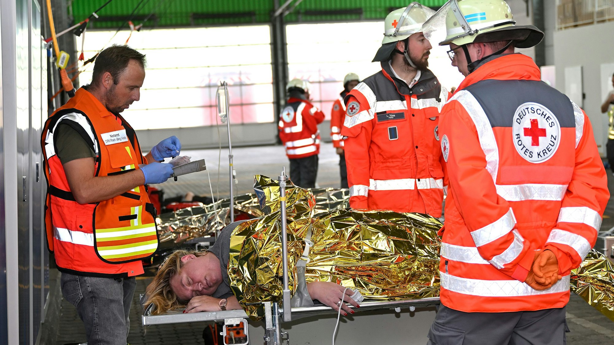 Ein Notarzt steht neben einer Trage, auf der eine Schwerverletzte eingewickelt in eine Rettungsdecke liegt. Zwei Rettungssanitäter stehen daneben.