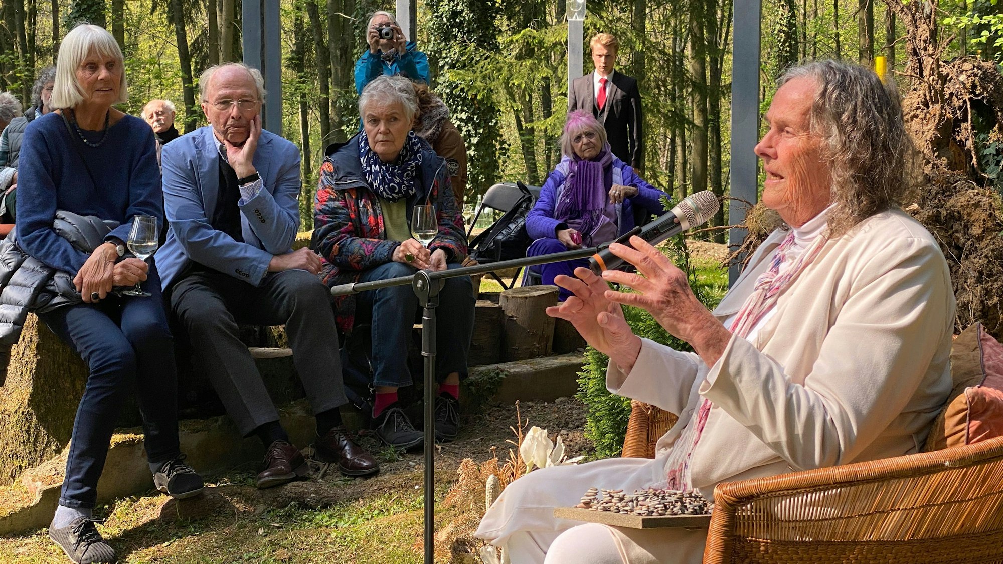 Künstlerin Mary Bauermeister sitzt in einem Korbstuhl und spricht, Menschen sitzen im Hintergrund und hören ihr zu.