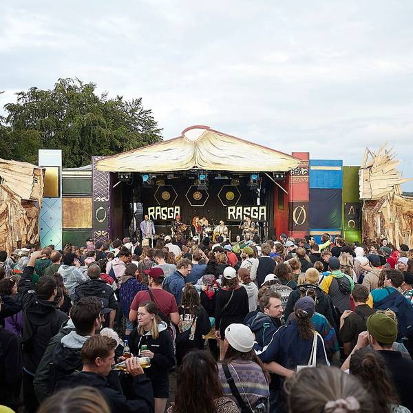 Das Bild zeigt zahlreiche Besucher vor einer Bühne auf einem Festivalgelände. Rechts und links stehen große Holzfiguren.