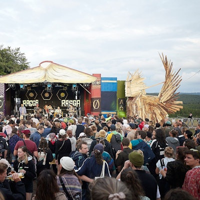 Das Bild zeigt zahlreiche Besucher vor einer Bühne auf einem Festivalgelände. Rechts und links stehen große Holzfiguren.