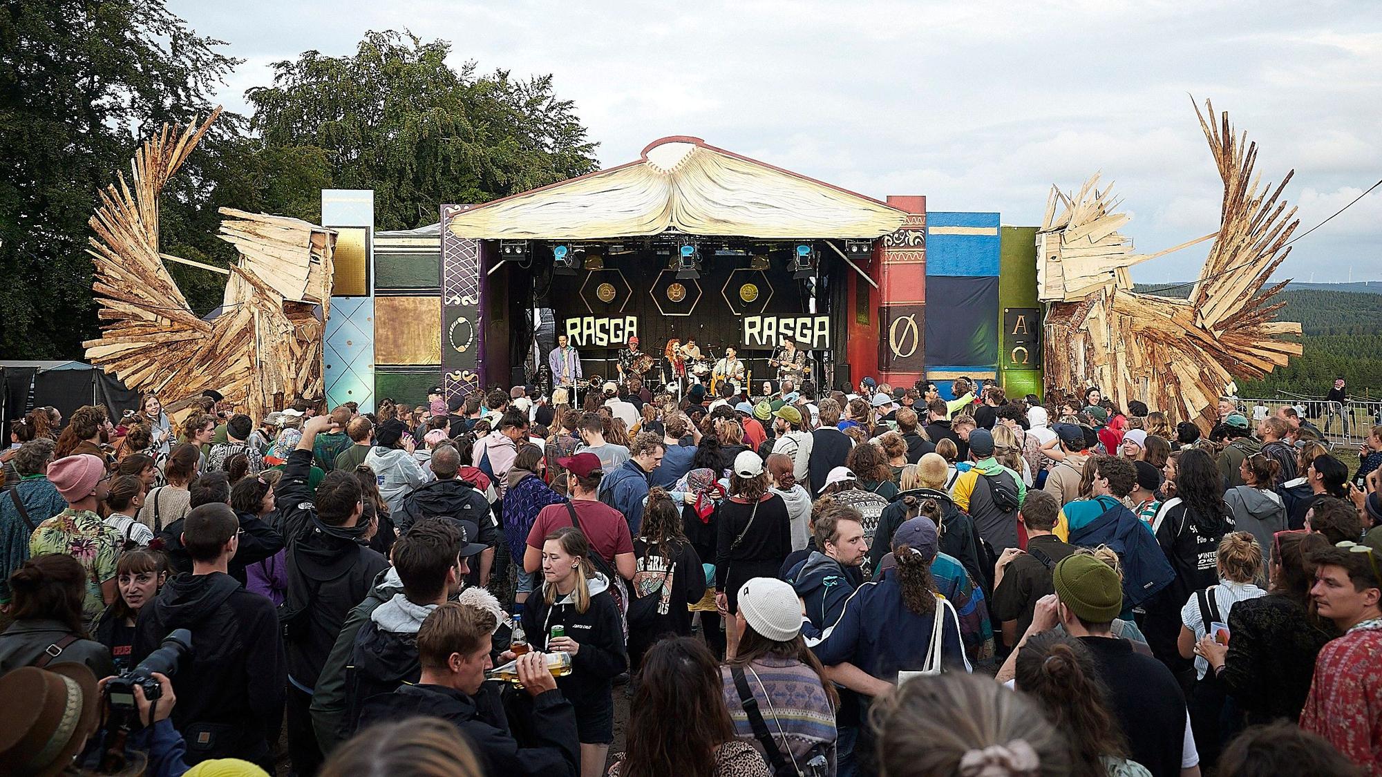 Das Bild zeigt zahlreiche Besucher vor einer Bühne auf einem Festivalgelände. Rechts und links stehen große Holzfiguren.