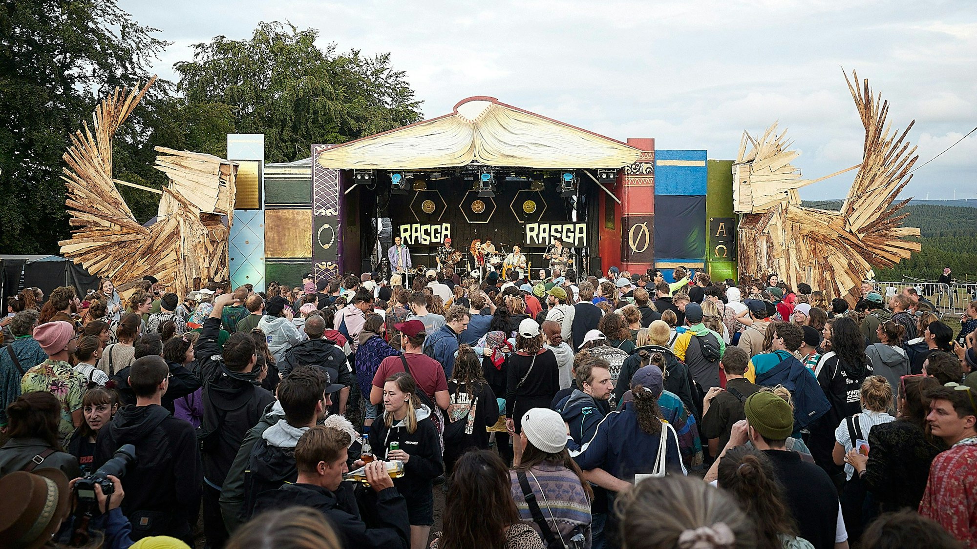 Das Bild zeigt zahlreiche Besucher vor einer Bühne auf einem Festivalgelände. Rechts und links stehen große Holzfiguren.