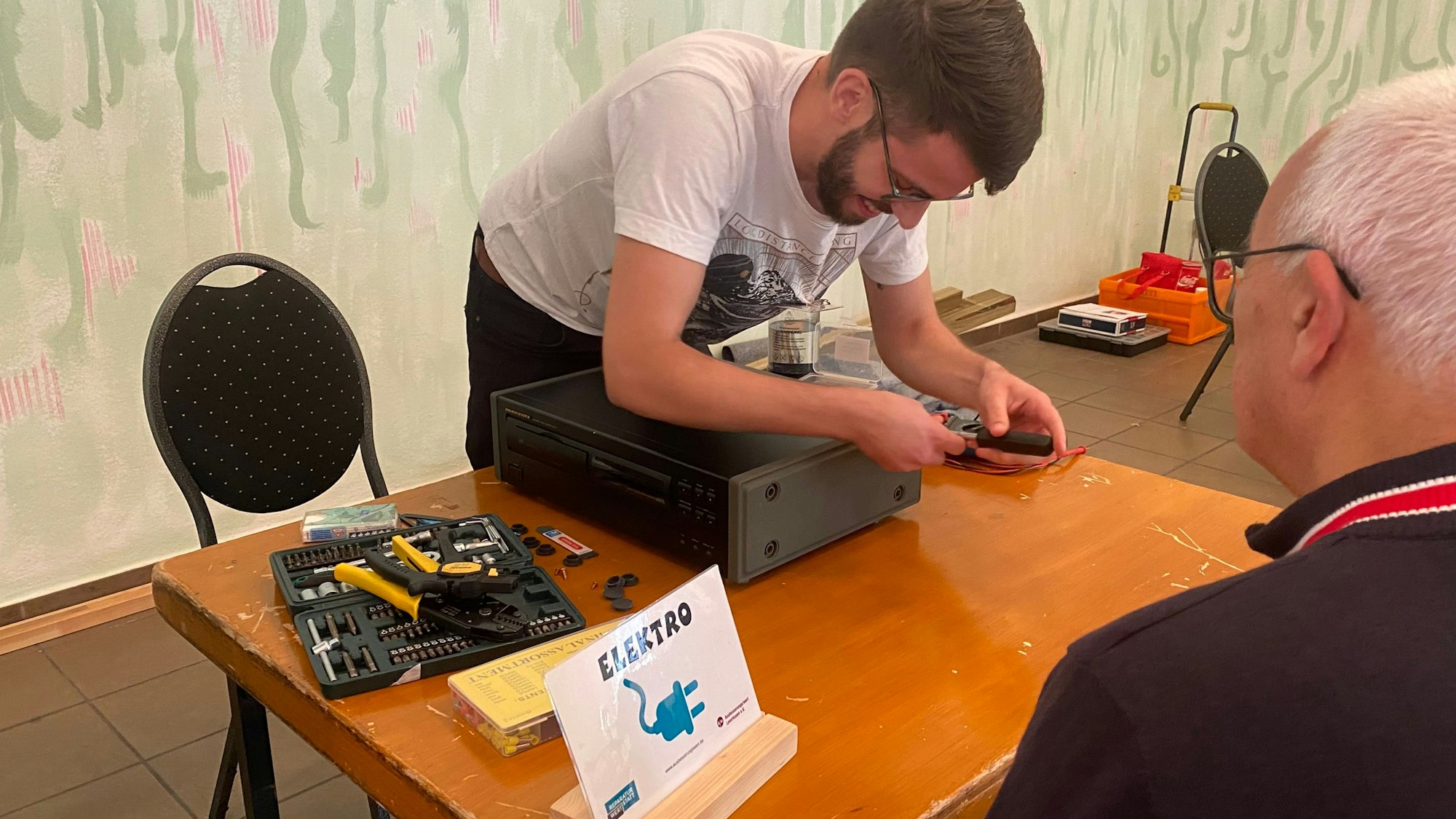 Manfred Hans steht vor einem Tisch, an dem ein Experte an seinem CD-Player schraubt.