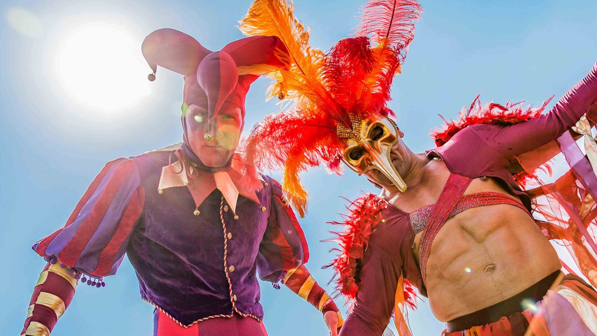 Bunt gekleidetete Animateure, mit Narrenhut und venizianischer Maske, stehen auf Stelzen und blicken in die Kamera.